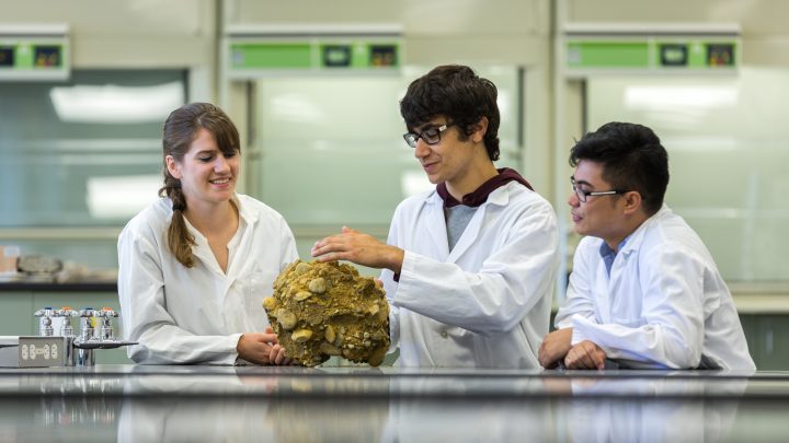 Three students in lab coats examining a large rock