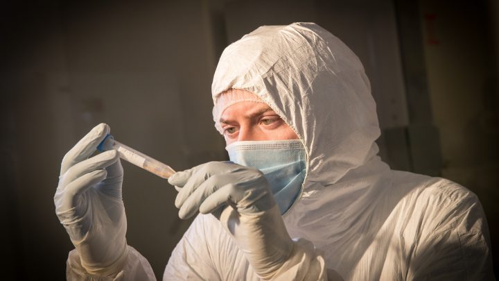 A man wearing personal protective equipment examining a vial