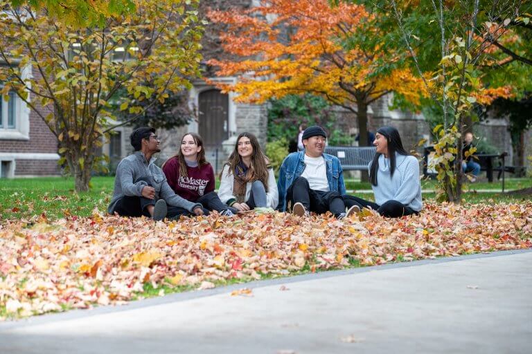 Five students sitting amongst a pile of fall leaves on campus