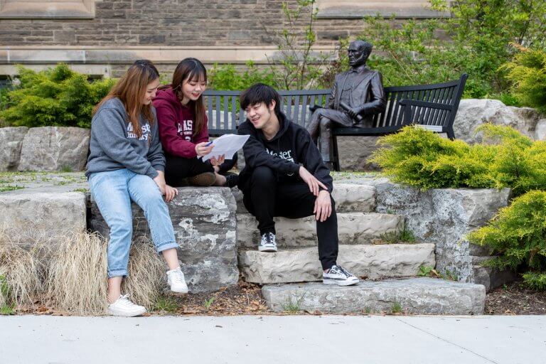 Three students sitting next to the William McMaster statue on campus, looking at a sheet of paper