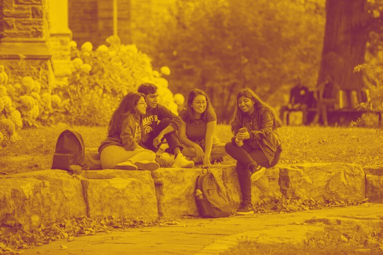 A maroon yellow duotone image of four students sitting together on large rocks on campus and talking