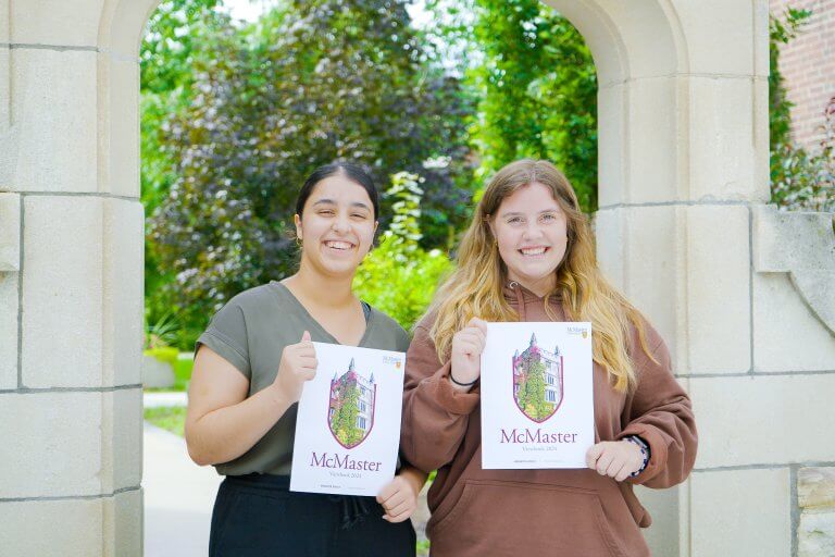 Two female tourguide and McMaster students are holding the latest McMaster viewbook in front of the arch.